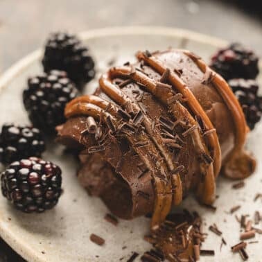 A scoop of chocolate cremeux topped with caramel sauce and chocolate shavings, served with fresh blackberries on a ceramic plate.