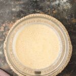 A glass bowl filled with a pale yellow, frothy liquid sits on a woven mat atop a dark, textured surface. A cloth is partially visible in the lower left corner.