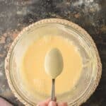 A hand holds a spoon above a glass bowl filled with pale yellow batter on a woven mat, set on a dark marbled surface.