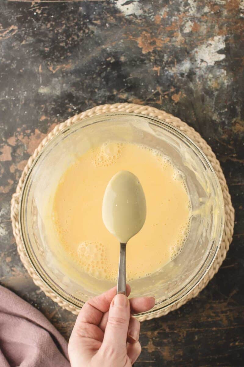 A hand holds a spoon above a glass bowl filled with pale yellow batter on a woven mat, set on a dark marbled surface.