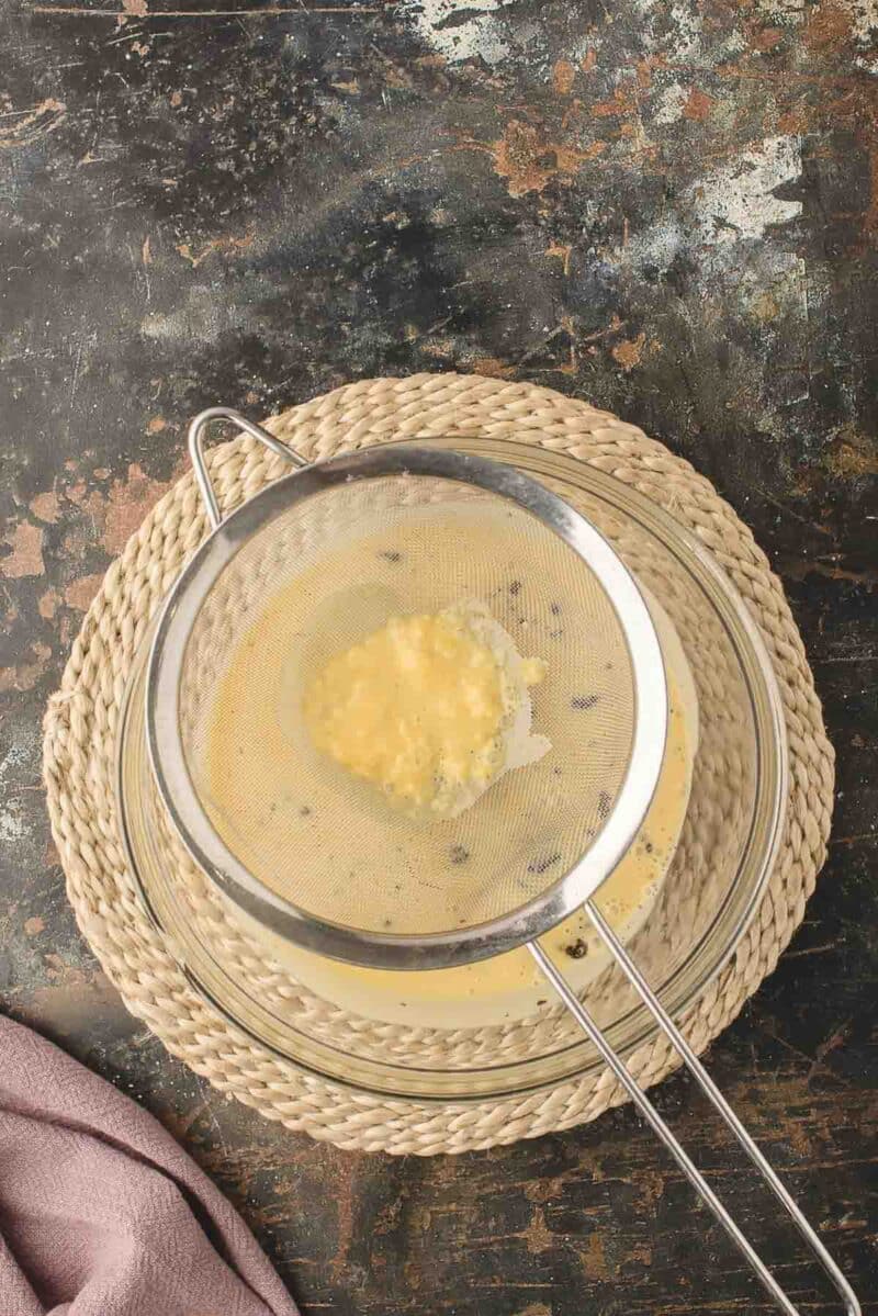 A metal strainer with yellow liquid and lumps sits over a glass bowl on a woven mat, set on a dark, textured surface.