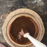 A glass bowl filled with chocolate batter being mixed with an immersion blender, placed on a woven mat against a dark, textured surface.