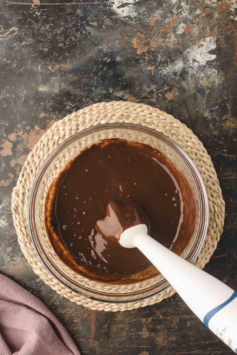 A glass bowl filled with chocolate batter being mixed with an immersion blender, placed on a woven mat against a dark, textured surface.