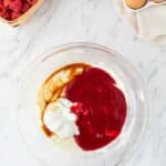 A glass bowl with yogurt, raspberry sauce, and vanilla extract on a marble counter, surrounded by raspberries, chocolate chips, and eggs.