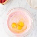 A glass bowl with pink batter and three cracked eggs, next to a basket of raspberries, a bowl of chocolate chips, and a beige cloth on a marble surface.