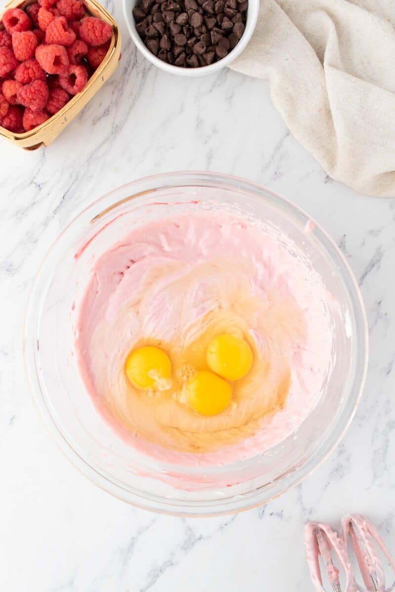 A glass bowl with pink batter and three cracked eggs, next to a basket of raspberries, a bowl of chocolate chips, and a beige cloth on a marble surface.