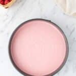 A round pan filled with smooth pink cheesecake batter sits on a marble surface, with a basket of raspberries and a bowl of chocolate chips nearby.