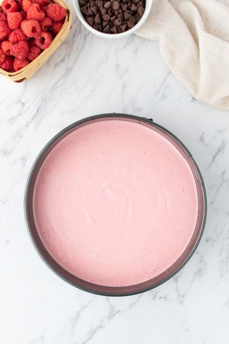 A round pan filled with smooth pink cheesecake batter sits on a marble surface, with a basket of raspberries and a bowl of chocolate chips nearby.