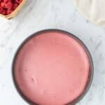 A round pan filled with smooth pink cheesecake sits on a marble surface, next to a basket of raspberries and a bowl of chocolate chips.