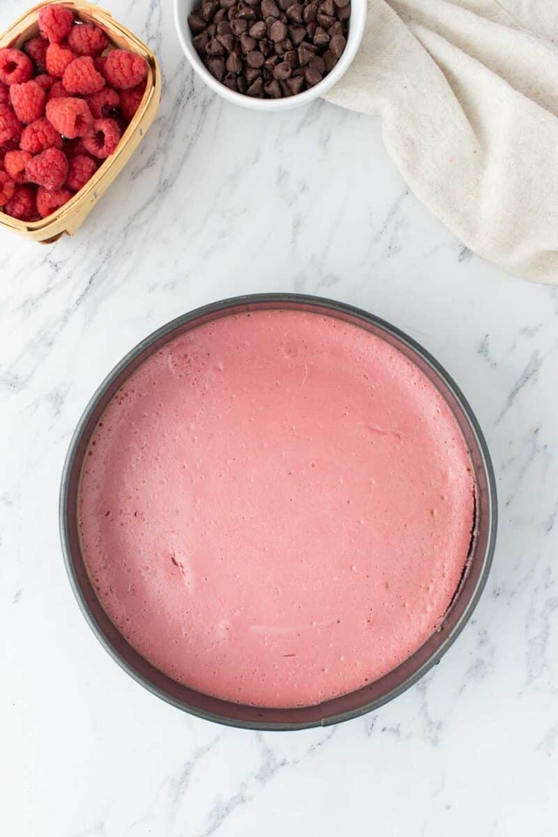 A round pan filled with smooth pink cheesecake sits on a marble surface, next to a basket of raspberries and a bowl of chocolate chips.