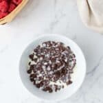 A white bowl filled with chocolate chips and cream sits on a marble countertop near a basket of raspberries and a beige cloth.
