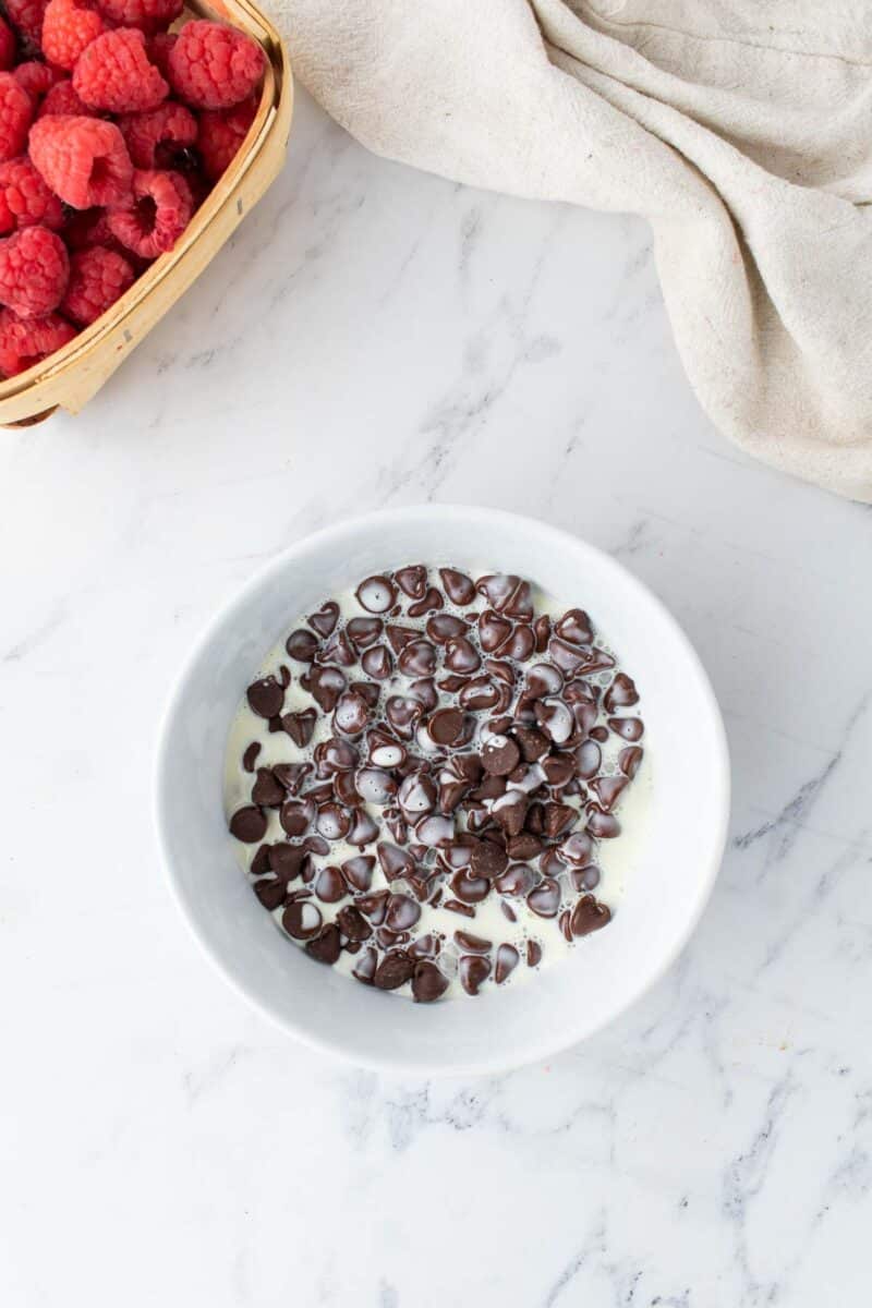 A white bowl filled with chocolate chips and cream sits on a marble countertop near a basket of raspberries and a beige cloth.