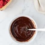 A bowl of smooth melted chocolate with a spoon, next to a basket of fresh raspberries on a marble surface.