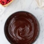 A round pan filled with smooth chocolate batter sits on a marble surface next to a small basket of fresh raspberries and a beige cloth.