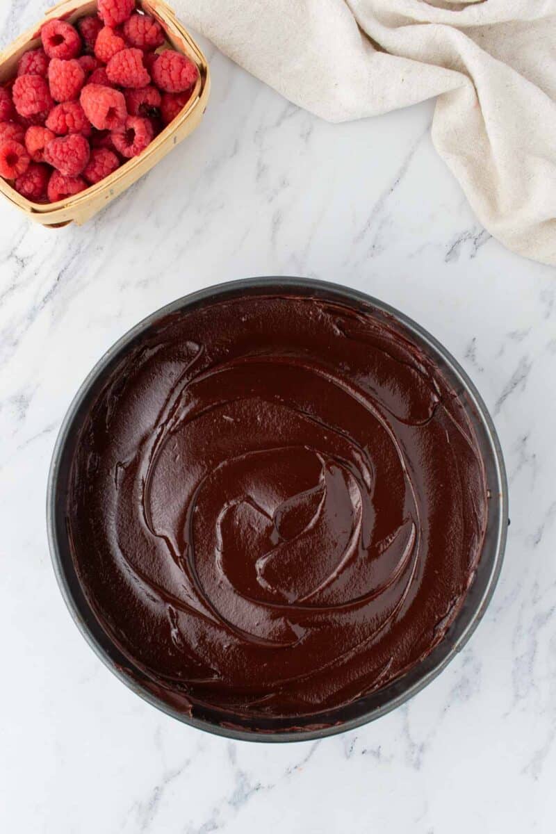 A round pan filled with smooth chocolate batter sits on a marble surface next to a small basket of fresh raspberries and a beige cloth.