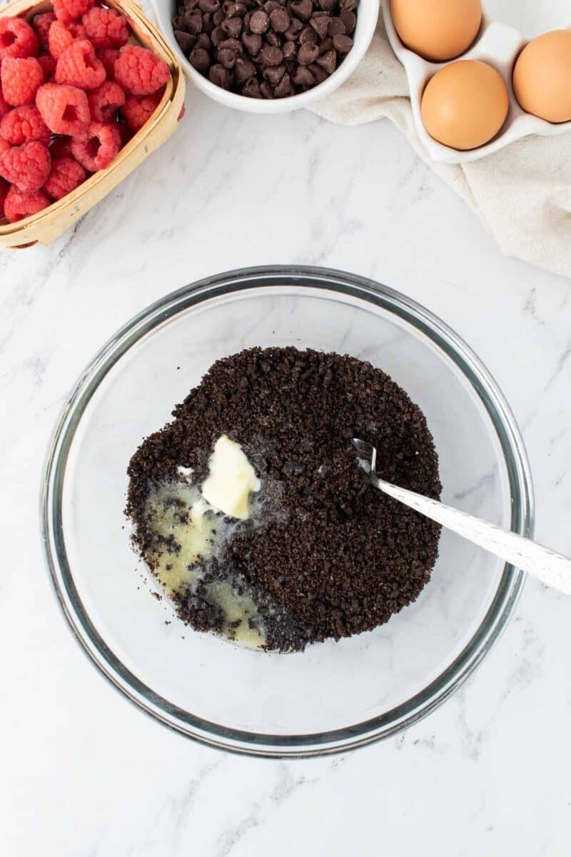 A glass bowl with crushed cookies and melted butter being mixed with a spoon, surrounded by eggs, raspberries, and chocolate chips on a marble countertop.