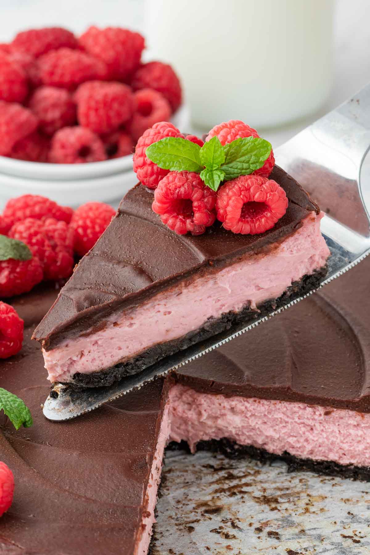 A slice of chocolate-covered raspberry cheesecake with an Oreo crust, topped with fresh raspberries and a mint leaf, is being lifted from the whole cake.