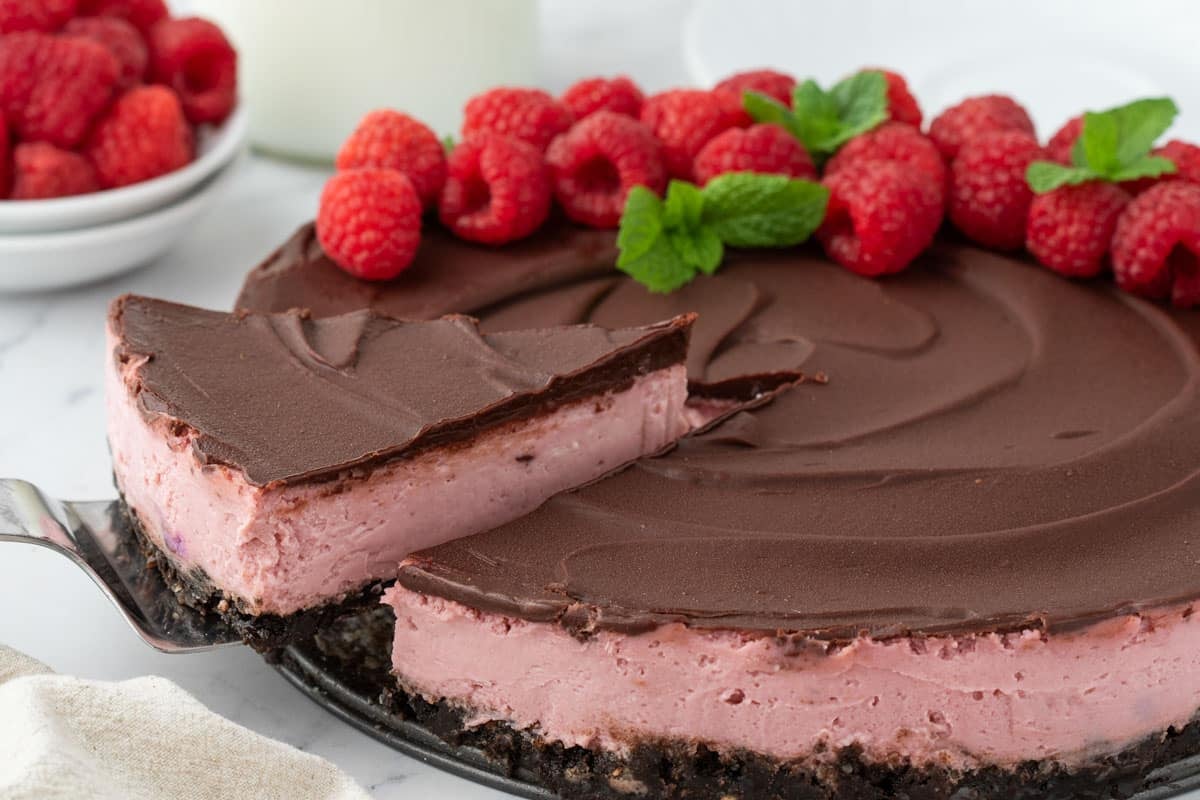 A chocolate-topped raspberry cheesecake is being sliced, garnished with fresh raspberries and mint leaves on top. A bowl of raspberries is visible in the background.