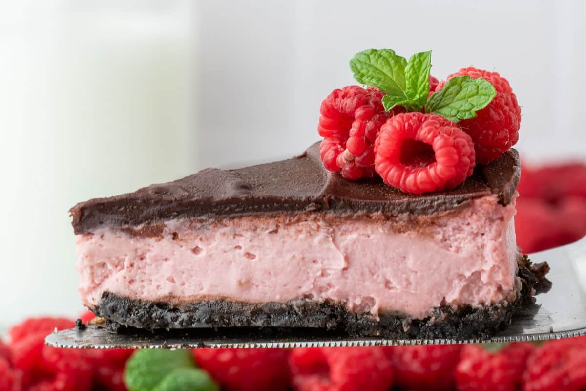 A slice of chocolate-topped raspberry cheesecake on an Oreo crust, garnished with fresh raspberries and a mint sprig.
