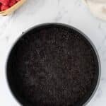 A round pan with a dark cookie crumb crust sits on a marble surface, surrounded by raspberries, chocolate chips, and eggs.
