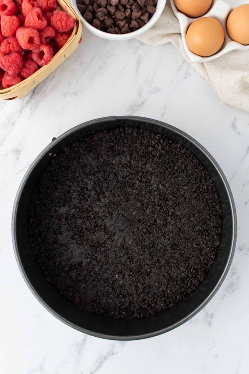 A round pan with a dark cookie crumb crust sits on a marble surface, surrounded by raspberries, chocolate chips, and eggs.