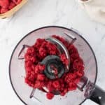 A food processor with raspberries inside sits on a marble counter next to a basket of raspberries, a bowl of chocolate chips, and three eggs.