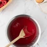 A saucepan of raspberry sauce, stirred with a wooden spoon, sits on a marble surface next to fresh raspberries, chocolate chips, and eggs.