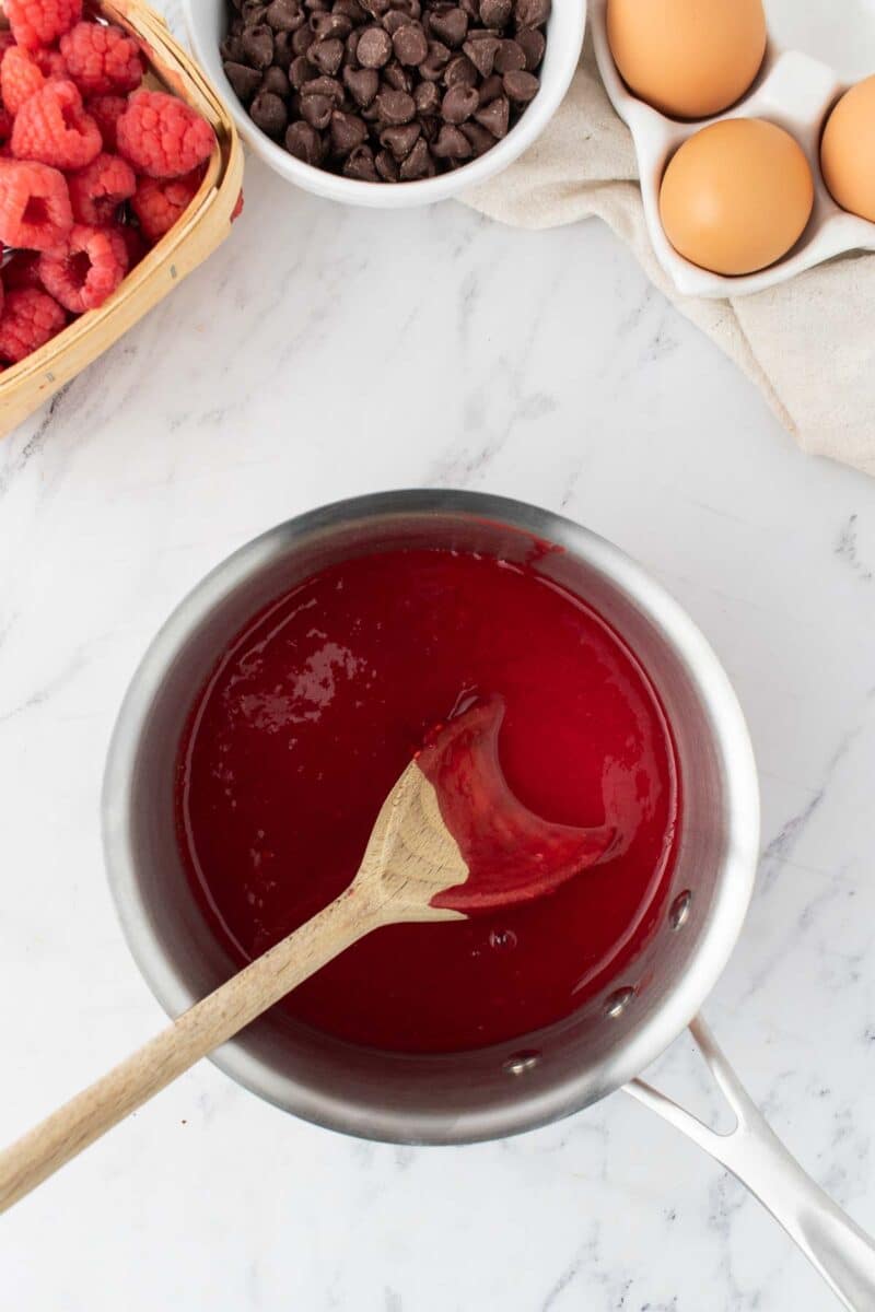 A saucepan of raspberry sauce, stirred with a wooden spoon, sits on a marble surface next to fresh raspberries, chocolate chips, and eggs.