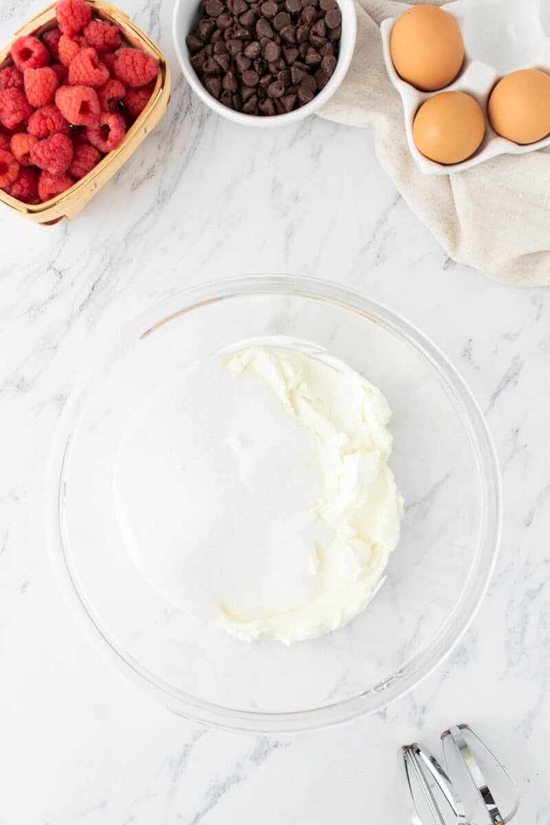 A glass bowl with cream cheese and sugar, surrounded by raspberries, chocolate chips, eggs, and a hand mixer on a marble countertop.