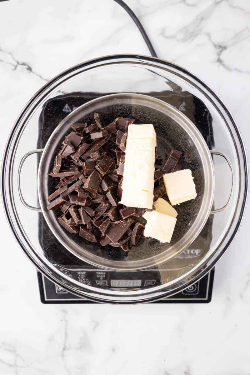 Chopped dark chocolate and blocks of butter in a metal bowl set over a pot of hot water on an electric stovetop, prepared for melting.
