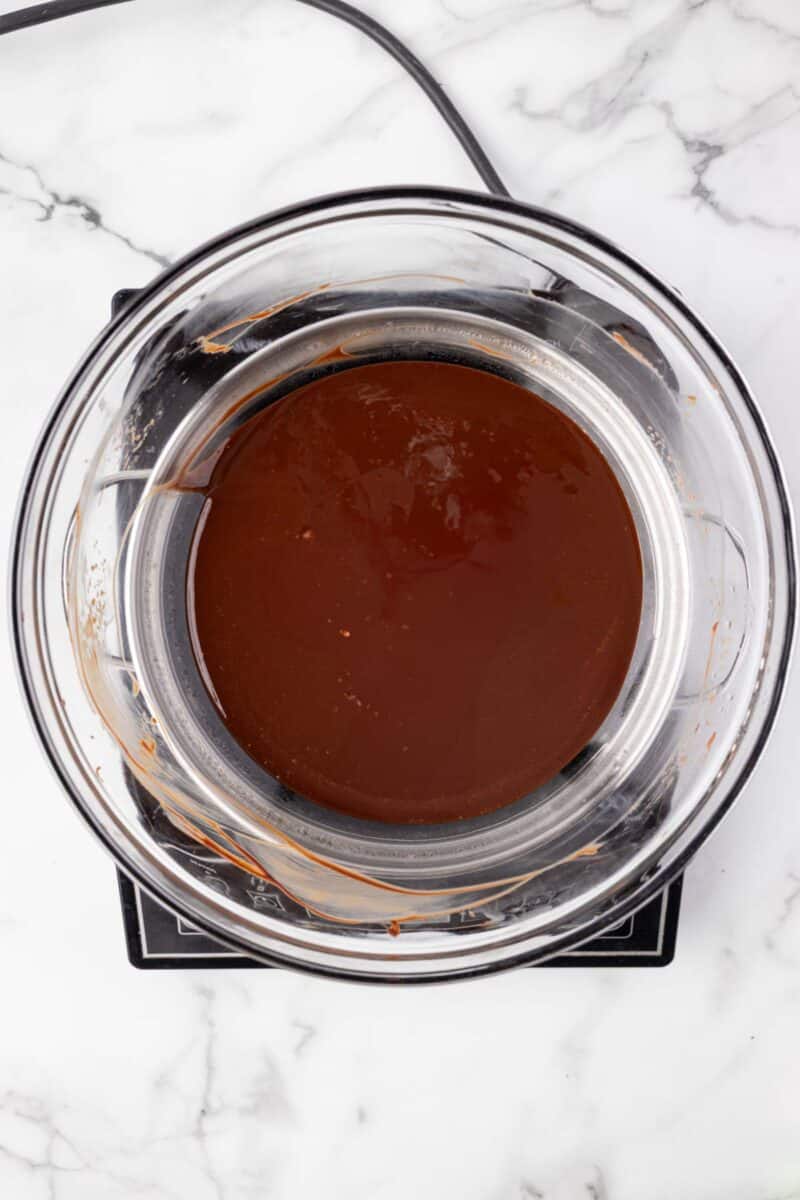 A glass bowl filled with melted chocolate sits over a saucepan on a marble countertop, viewed from above.
