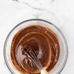 A hand is whisking chocolate batter in a clear glass bowl on a white marble surface.