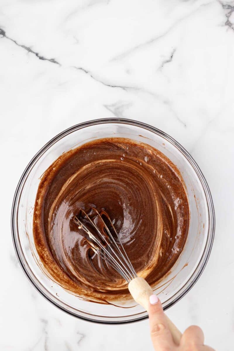 A hand is whisking chocolate batter in a clear glass bowl on a white marble surface.