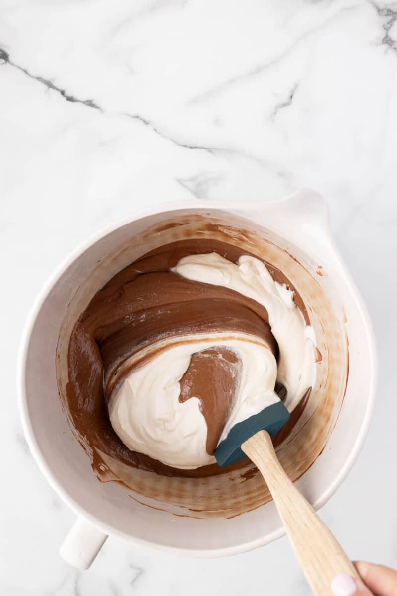 A mixing bowl with chocolate and white batter being folded together with a spatula on a marble surface.