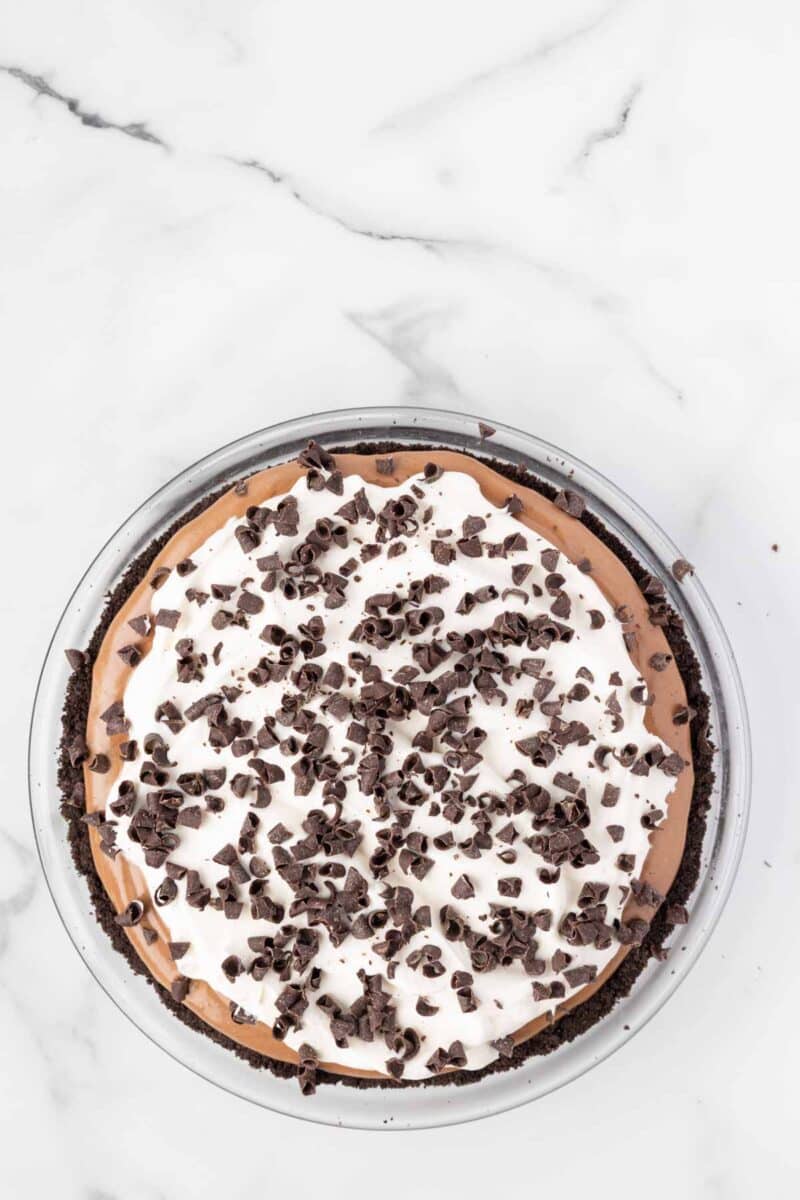 A chocolate cream pie with a whipped topping and chocolate chips, displayed in a glass pie dish on a white marble surface.