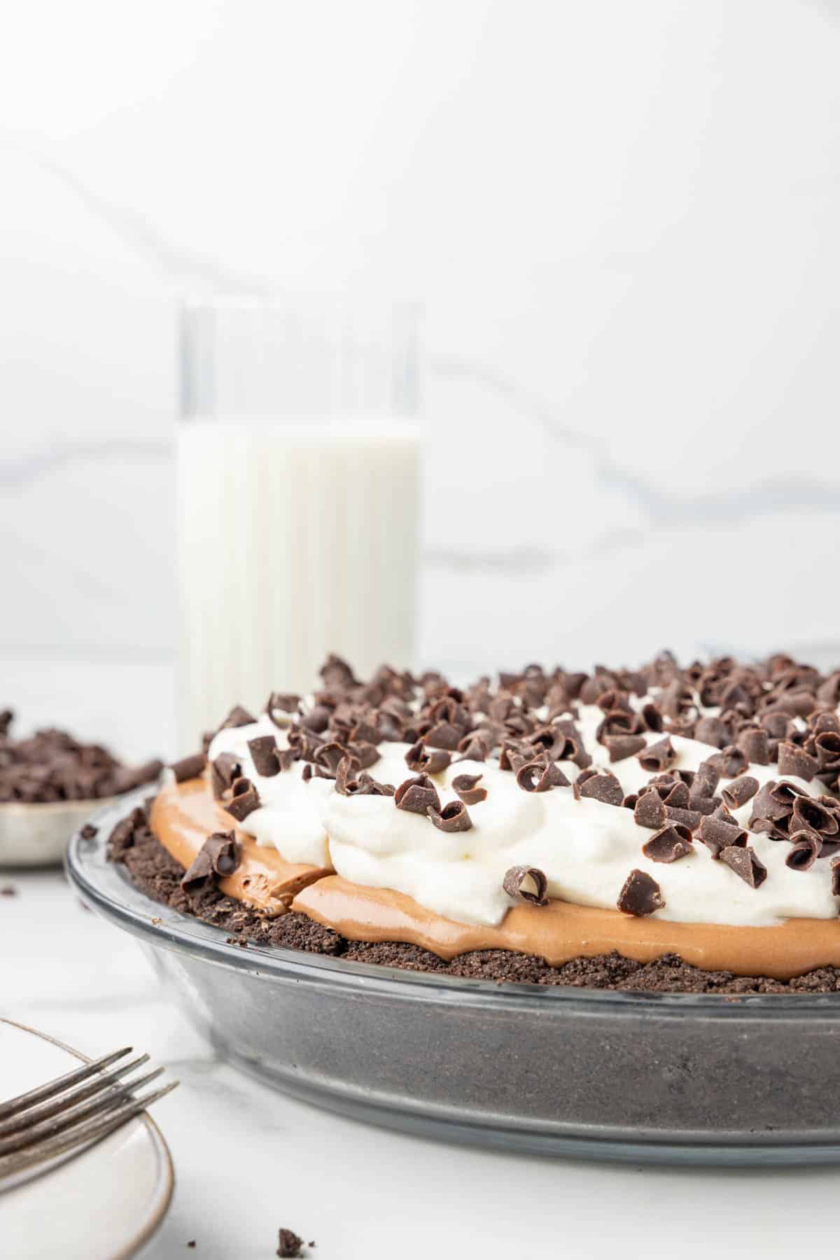 A chocolate cream pie with whipped topping and chocolate shavings in a glass pie dish, with a glass of milk in the background.