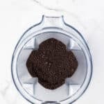 A top-down view of finely ground dark powder, likely coffee or cookie crumbs, inside a clear blender container on a white marble surface.