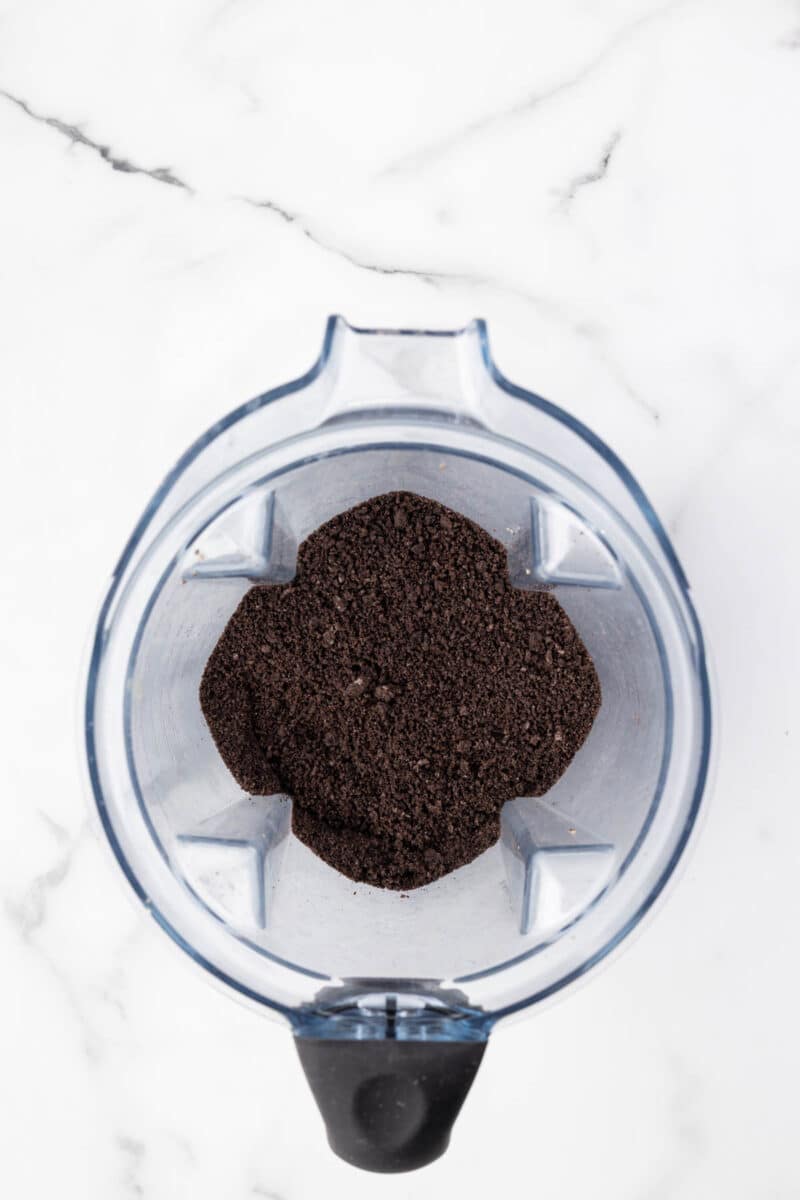 A top-down view of finely ground dark powder, likely coffee or cookie crumbs, inside a clear blender container on a white marble surface.