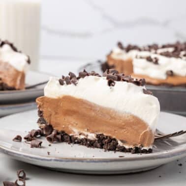 A slice of chocolate cream pie with whipped topping and chocolate chips on a plate, with the rest of the pie and a glass of milk in the background.