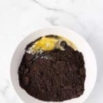 A white bowl with crushed chocolate cookies and a cracked egg on a white marble surface.