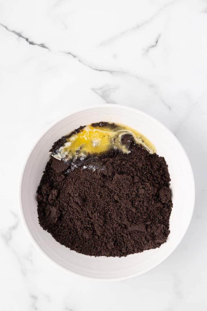 A white bowl with crushed chocolate cookies and a cracked egg on a white marble surface.