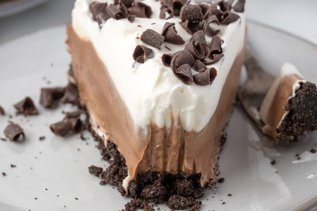 A slice of chocolate and cream layered pie with chocolate shavings on top and a crumbly chocolate crust, served on a white plate.