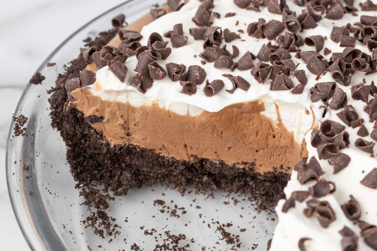 A chocolate cream pie with a slice removed, showing layers of chocolate filling, whipped topping, chocolate curls, and an Oreo cookie crust.