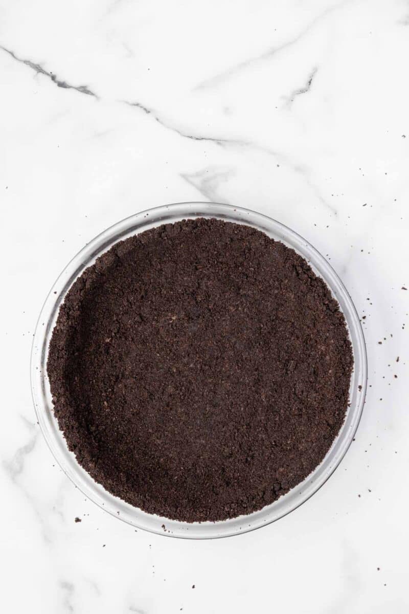 A glass pie dish filled with a pressed chocolate cookie crumb crust on a white marble surface.