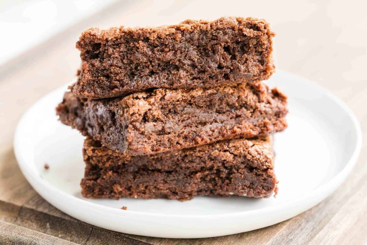 Three chocolate brownies are stacked on a white plate placed on a wooden surface.