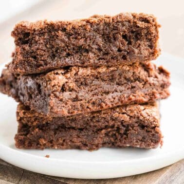 Three chocolate brownies are stacked on a white plate placed on a wooden surface.