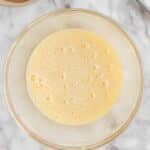 A glass bowl filled with a beaten egg mixture sits on a marble surface next to a whisk, a bowl of cocoa powder, and a striped cloth.