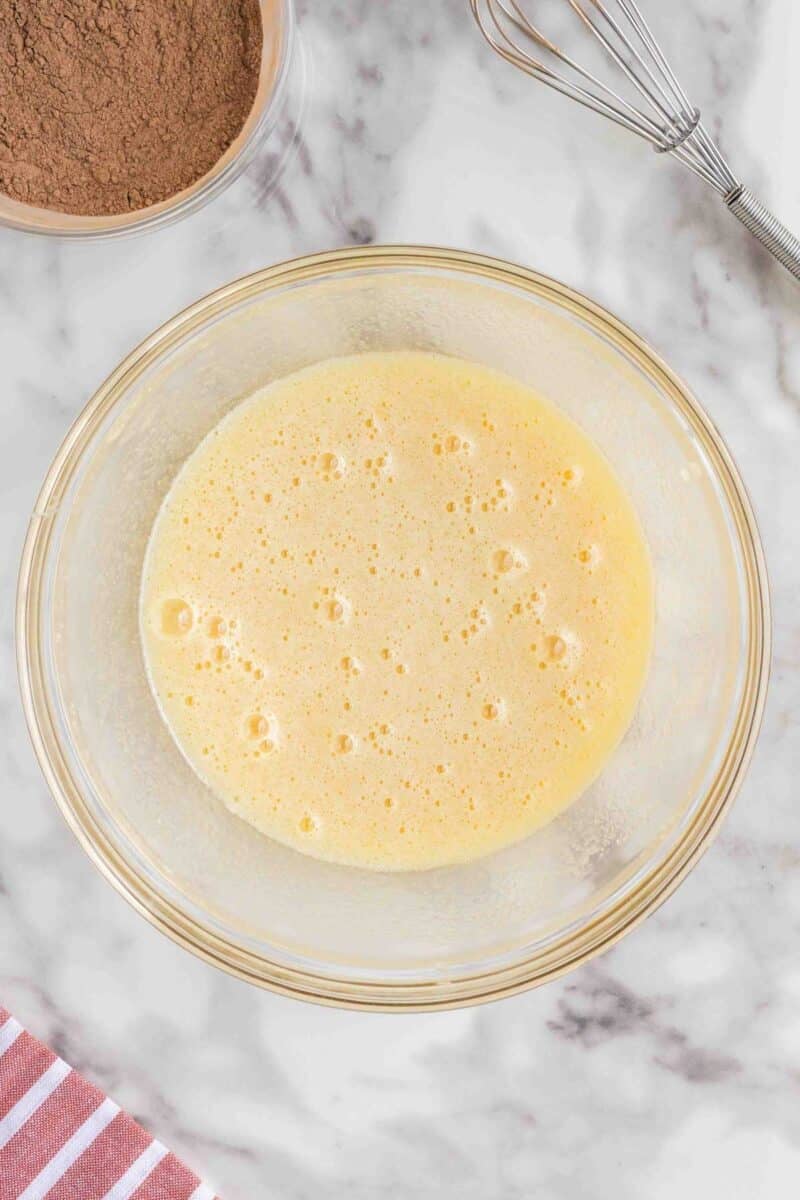 A glass bowl filled with a beaten egg mixture sits on a marble surface next to a whisk, a bowl of cocoa powder, and a striped cloth.