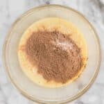 A glass bowl with a mixture of beaten eggs and cocoa powder on a marble surface, with a whisk and striped towel nearby.