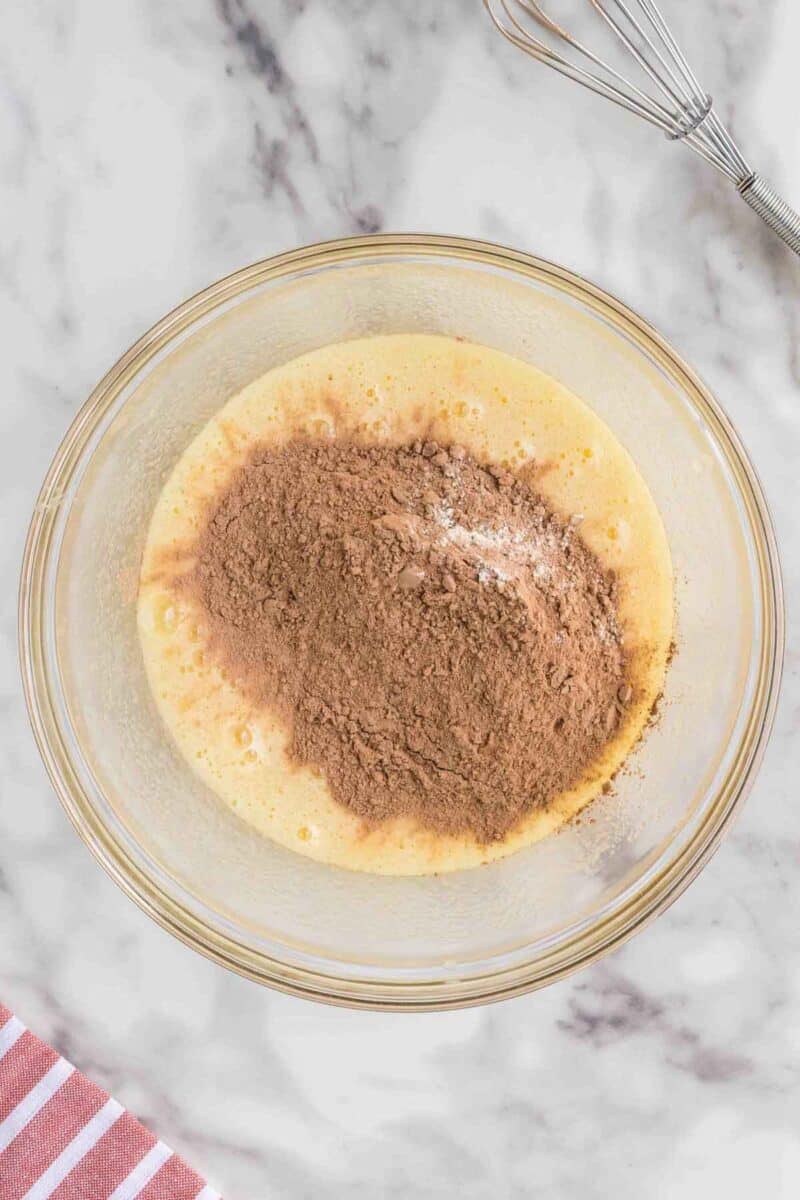 A glass bowl with a mixture of beaten eggs and cocoa powder on a marble surface, with a whisk and striped towel nearby.